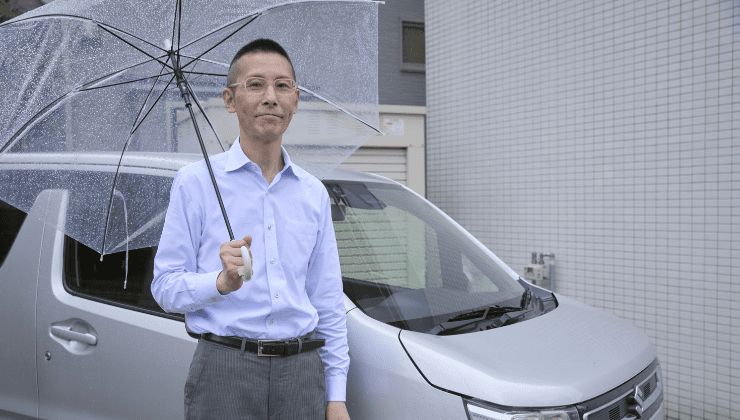 ワゴンRの前で傘をさして立つ薬局経営者