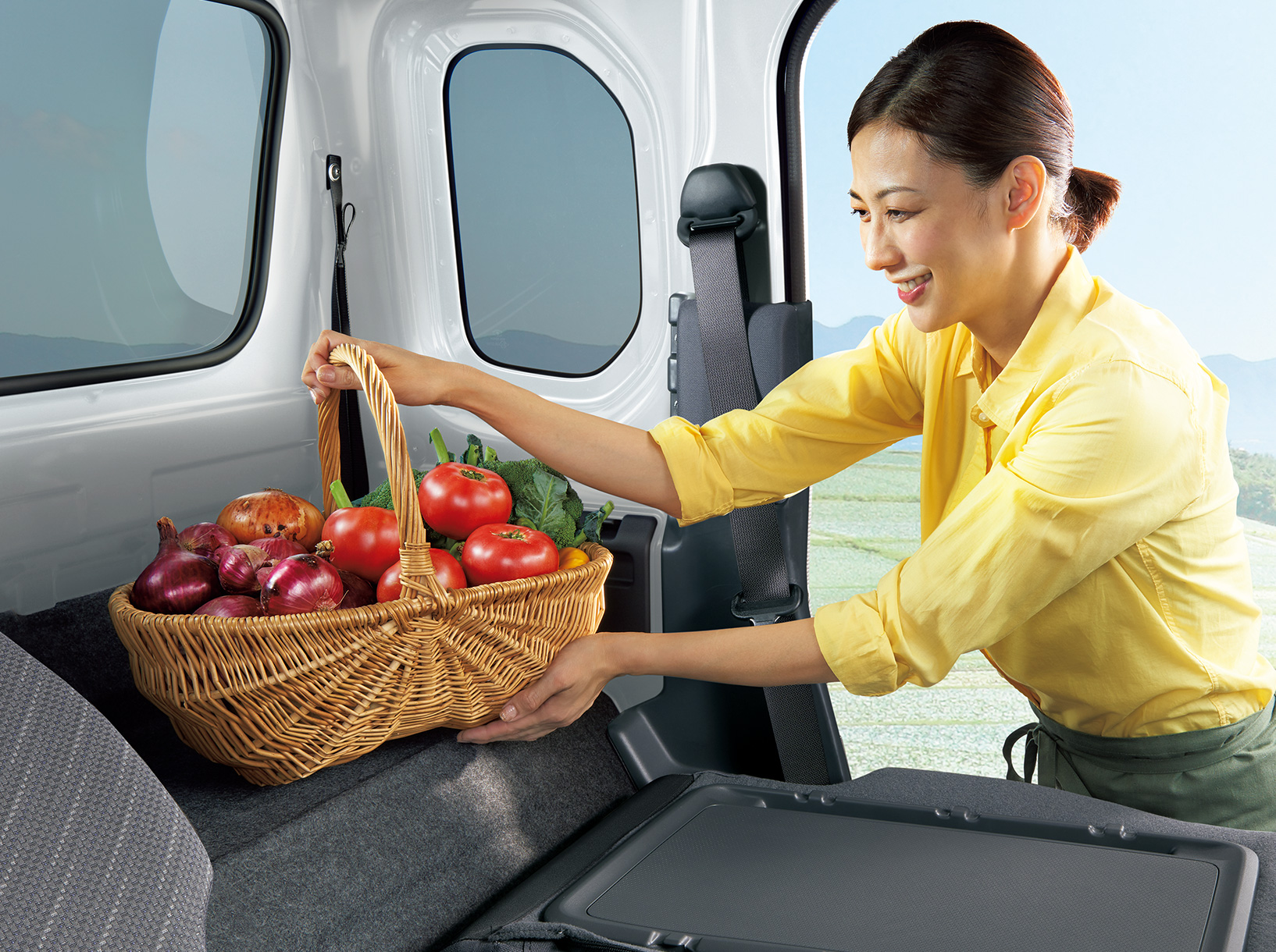 女性が野菜の入ったかごをスーパーキャリイのシートバックスペースに載せている様子。スーパーキャリイ X 5MT車、ボディーカラーはホワイト（26U）。